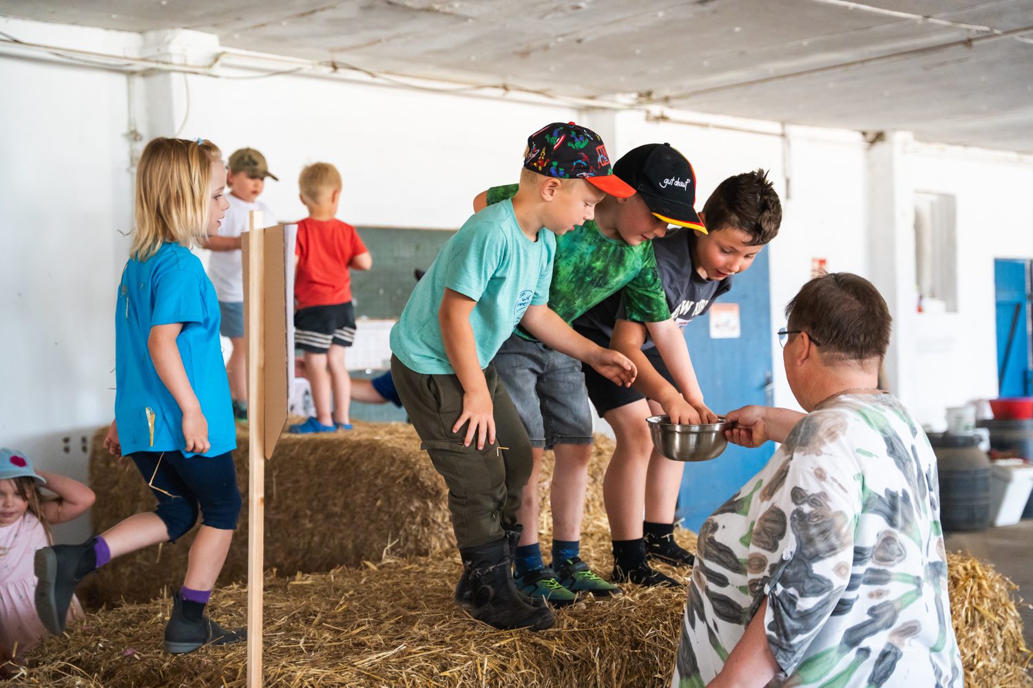 Kinder stehen in der Strohhalle auf Strohballen und schauen in eine Schüssel, die eine Betreuerin hält