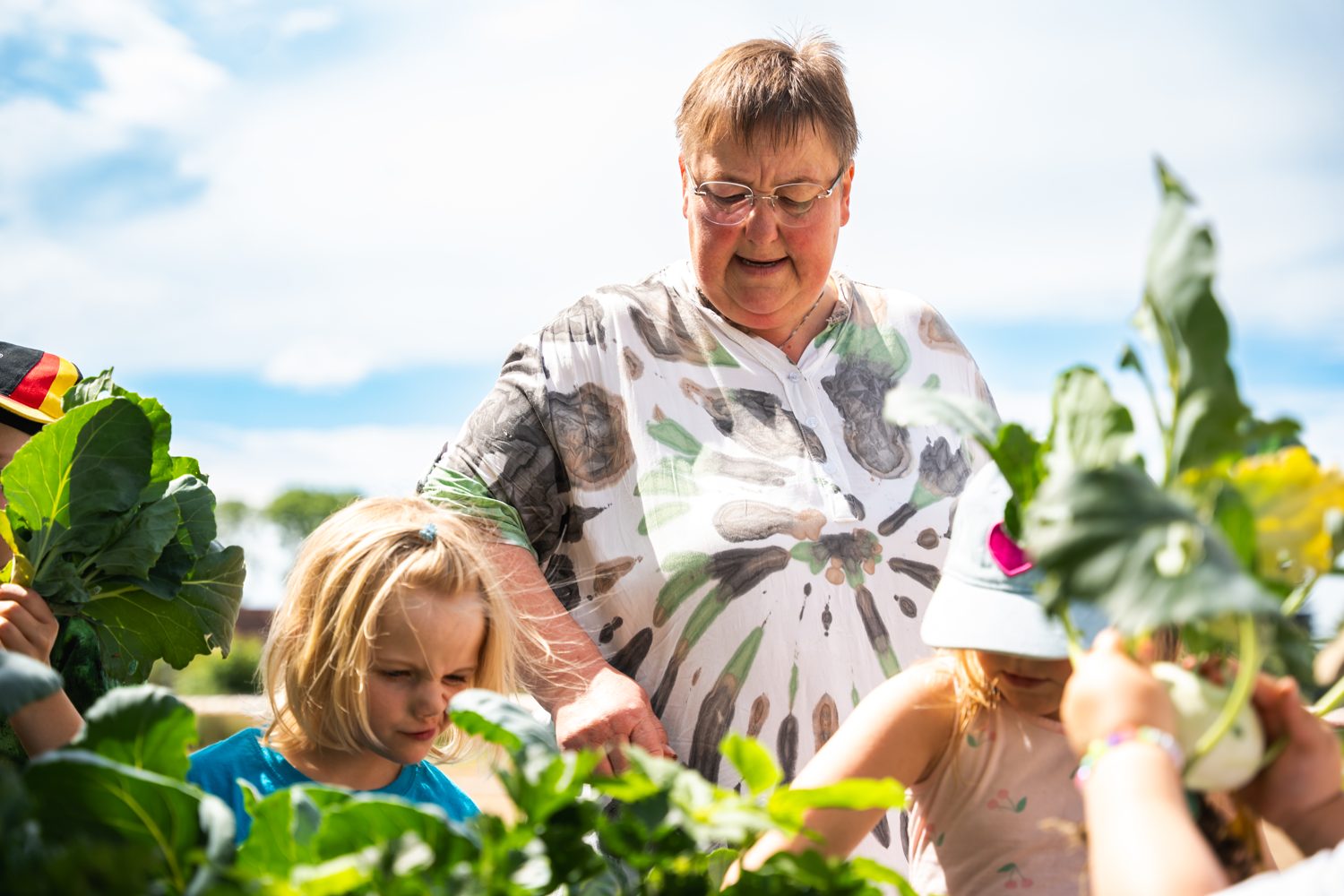 Eine Frau erntet mit Kindern Kohlrabi im Hochbeet unter blauem Himmel